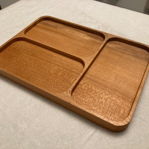 Puede incluir: Bandeja rectangular de madera para servir con tres compartimentos. La bandeja está hecha de madera de color marrón claro con un patrón de vetas natural. Los compartimentos están separados por divisores curvos y la bandeja tiene bordes redondeados.