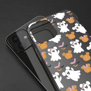 May include: A clear phone case with a Halloween-themed pattern. The design features white ghosts with pink bows, orange pumpkins, and pink bats on a dark gray background. The case is shown on top of a black smartphone.