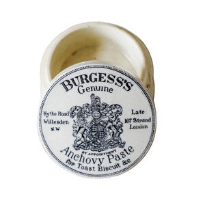 May include: An open, cream-coloured ceramic jar with a lid. The lid is printed with the text "BURGESS'S Genuine Anchovy Paste", a crest, and the product's location. The jar is set against a white background.
