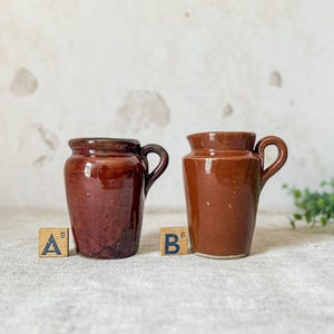 May include: Two antique brown ceramic jars with handles. The jar on the left is a darker shade of brown with a textured surface. The jar on the right is a lighter shade of brown with a smooth surface. Both jars have a rustic, vintage aesthetic.