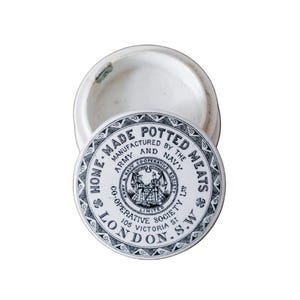 May include: An antique white ceramic jar with a lid. The lid features a black and white circular design with text that reads "HOME MADE POTTED MEATS" and "LONDON S.W.". The jar is open, revealing its interior.