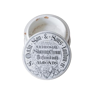 May include: An antique white ceramic shaving cream jar with a lid. The lid features black text that reads "Dam Son & Sons, London, er." and "Ambrosial Shaving Cream Perfumed with Almonds." The jar has a distressed appearance.