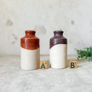 May include: Two small, vintage ceramic bottles. One is brown and cream, the other is dark brown and cream. The bottles are cylindrical with a rounded top. The letters "A" and "B" are on small wooden blocks.
