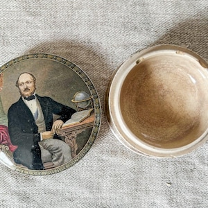 May include: An open, round, ceramic trinket box with a lid featuring a portrait of a man in a black suit. The lid's border has a decorative pattern. The text on the lid reads "THE LATE PRINCE CONSORT".