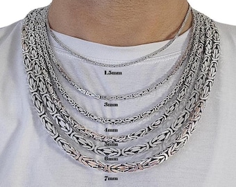 May include: A collection of silver-colored Byzantine chain necklaces in various widths, ranging from 1.5mm to 7mm, displayed on a person wearing a white t-shirt. The necklaces are arranged from thinnest to thickest.