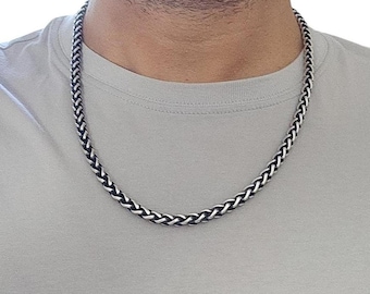 May include: A silver and black braided chain necklace. The necklace is made of interwoven links, creating a textured, rope-like appearance. The chain is worn around the neck, resting on a gray shirt.