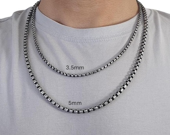 May include: Two silver-colored box chain necklaces, 3.5mm and 5mm, are displayed. The necklaces are worn around the neck. The image shows the difference in size between the two chains. The background is a light gray t-shirt.