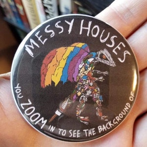 May include: A black and white button with the text "Messy Houses You Zoom In To See The Background Of" in white lettering. The button features a colourful rainbow-like design with a cartoon figure in the centre.