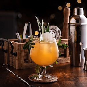 K&ouml;nnte beinhalten: Ein gelber Cocktail in einem Stielglas mit einem gro&szlig;en Eisw&uuml;rfel und einem Zweig gr&uuml;ner Bl&auml;tter. Das Getr&auml;nk ist mit einem Ananasst&uuml;ck und einem Minzzweig garniert. Das Glas steht auf einem geflochtenen Untersetzer auf einem Holztisch.