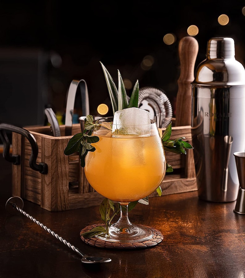 K&ouml;nnte beinhalten: Ein gelber Cocktail in einem Stielglas mit einem gro&szlig;en Eisw&uuml;rfel und einem Zweig gr&uuml;ner Bl&auml;tter. Das Getr&auml;nk ist mit einem Ananasst&uuml;ck und einem Minzzweig garniert. Das Glas steht auf einem geflochtenen Untersetzer auf einem Holztisch.
