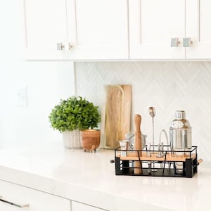 May include: A black metal barware rack with a wooden base holds a cocktail shaker, a muddler, a jigger, and other bar tools. The rack is on a white countertop in a kitchen.