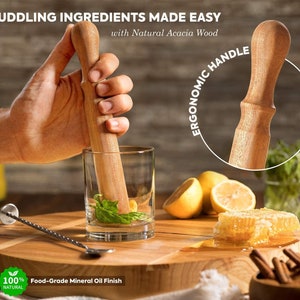 May include: A wooden muddler with an ergonomic handle, made from natural acacia wood. The muddler is being used to crush ingredients in a glass with a clear liquid and green herbs. The muddler has a food-grade mineral oil finish.