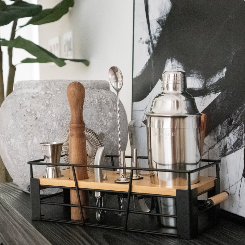 May include: A black metal wire basket with a wooden handle and a wooden bottom. The basket contains a variety of bar tools, including a cocktail shaker, a jigger, a muddler, a bar spoon, and a strainer.