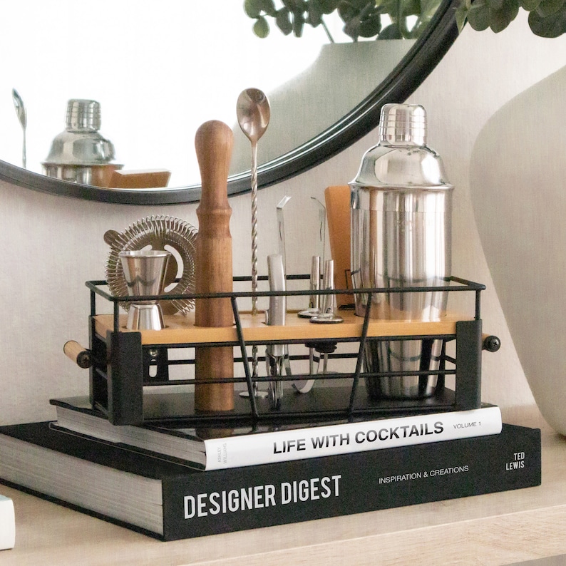 May include: A black metal bar cart organizer with a wooden shelf, holding a variety of bar tools, including a cocktail shaker, jigger, muddler, and bar spoon. The organizer is sitting on top of two books, one titled "Designer Digest" and the other titled "Life with Cocktails".