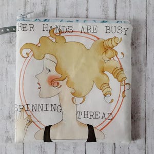 Könnte beinhalten: Eine weiße Stofftasche mit Reißverschluss und einem blau-weiß gestreiften Rand. Die Tasche zeigt eine Aquarell-Illustration einer Frau mit blonden Haaren und dem Text "Her hands are busy spinning thread".