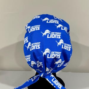 May include: A blue and white patterned fabric head wrap with the Detroit Lions logo repeated throughout. The wrap is tied at the back of the head with two long ties.
