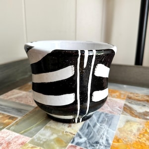 May include: A ceramic cup featuring a glossy black glaze with hand-painted white horizontal stripes and a single vertical white line running down the centre. The interior of the cup is finished in a smooth white glaze, creating a high-contrast look.