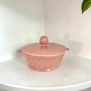 May include: A dusty rose ceramic serving bowl with a matching lid featuring a spherical knob handle. The bowl has a wide, circular base, a tapered body with subtle diagonal ribbed detailing, and two small, integrated side handles.