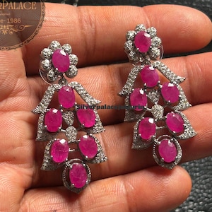 以下が含まれることがあります： 鮮やかなピンク色の楕円形宝石をあしらった、シルバーカラーのシャンデリアピアス。ピアスは上部に花のモチーフがあり、宝石が滝のように配置されています。Silverpalaceのロゴが見えます。