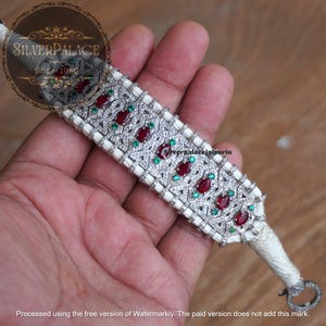 Op de afbeelding: Een zilveren armband met een wit koord, met een centrale band versierd met rode en groene edelstenen. De band heeft een geometrisch ontwerp met kleine, sprankelende accenten. De armband wordt in een hand gehouden.