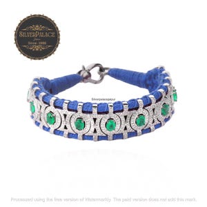 May include: A silver bracelet with a blue cord and a row of emerald green gemstones surrounded by diamonds. The bracelet has a silver clasp.