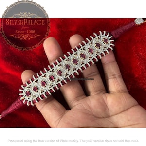 Puede incluir: Un brazalete de plata y rubíes con base de cordón rojo. El brazalete presenta rubíes ovalados engastados en un diseño de filigrana plateada, con pequeñas piedras transparentes. El texto "SILVERPALACE Jaipur Since-1986" es visible.