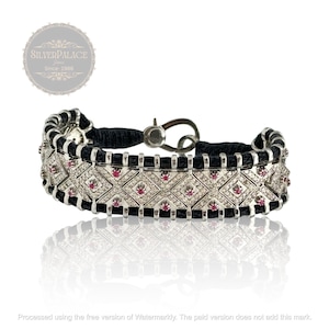 May include: A silver bracelet with a black leather strap and a geometric design featuring small white diamonds and pink gemstones. The bracelet has a silver clasp.
