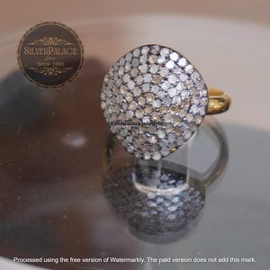 Puede incluir: Un anillo de oro con una gran cara redonda cubierta de pequeñas piedras brillantes. El anillo se presenta sobre una superficie reflectante, mostrando su intrincado diseño y los reflejos de la luz en las piedras. De Silver Palace.