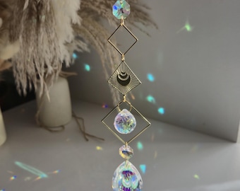 May include: A gold-colored suncatcher with a chain, featuring a faceted crystal, two square frames, and a teardrop-shaped crystal. The design includes a crescent moon and sunburst pattern. The crystals create a rainbow effect.