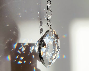 May include: A clear, faceted crystal prism, suspended by a silver chain. The prism is elongated with pointed ends. Light refracts through the crystal, creating a rainbow effect on the white background. The crystal is a decorative item.