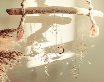 May include: A driftwood branch with six hanging crystal suncatchers. Each suncatcher is a different shape, including a circle, a star, a crescent moon, and a teardrop. The suncatchers are made of gold metal and have clear crystals that catch the light.