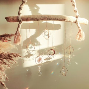 May include: A driftwood branch with six hanging crystal suncatchers. Each suncatcher is a different shape, including a circle, a star, a crescent moon, and a teardrop. The suncatchers are made of gold metal and have clear crystals that catch the light.