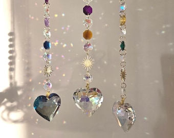 May include: Three crystal suncatchers with heart-shaped crystals hanging from gold chains. The chains are adorned with small, colorful crystals. The heart-shaped crystals are clear, iridescent, and reflect light.