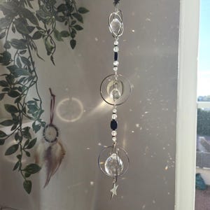 Peut inclure: Un attrape-soleil en cristal avec une étoile, des perles et des orbes de cristal suspendus à une chaîne argentée. L'attrape-soleil est suspendu devant une fenêtre.