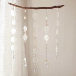 May include: A white, iridescent shell mobile with five strands of shells and crystals hanging from a driftwood branch. Each strand features a different combination of white, iridescent shells and clear crystals.