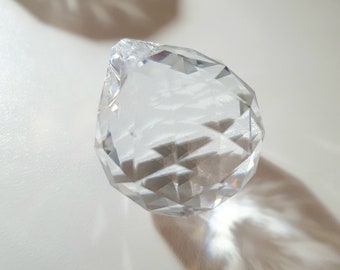Puede incluir: Un prisma de cristal de vidrio transparente con un diseño facetado. El prisma proyecta una sombra sobre una superficie blanca.