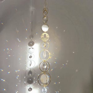 Puede incluir: Un atrapasol de cristal dorado con múltiples cuentas de cristal transparente y orbes de vidrio iridiscente blanco que cuelgan de una cadena. El atrapasol está colgado de un techo blanco.