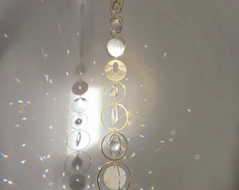 May include: A gold crystal suncatcher with multiple clear crystal beads and white iridescent glass orbs hanging from a chain. The suncatcher is hanging from a white ceiling.