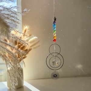 Puede incluir: Un atrapasoles colgante con una cadena plateada, que presenta una serie de cuentas de colores en un espectro de arcoíris. Debajo de las cuentas hay tres anillos de metal negro, un cristal facetado y un diseño de sol. El atrapasoles está suspendido frente a una pared blanca.