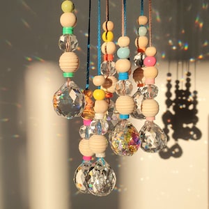 May include: A set of six crystal suncatchers hanging from wooden beads with colourful accents. Each suncatcher is a different shape and size, and they are all strung together on a long cord.