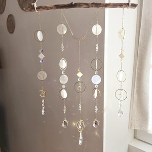 Peut inclure: Une suspension murale en macramé blanche et dorée avec de multiples brins de perles, de coquillages et de cristaux. Les brins sont attachés à une branche de bois naturel.