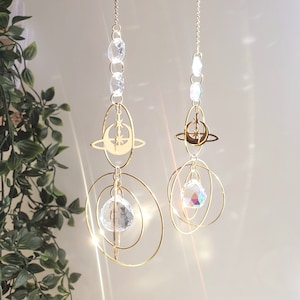 May include: Two gold celestial suncatchers with crystal prisms hanging from a chain. Each suncatcher features a crescent moon and star design with a circle around it. The suncatchers are suspended from a chain with clear crystal beads.