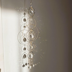 Puede incluir: Móvil decorativo colgante con anillos dorados, esferas de cristal transparente y detalles de estrellas. El móvil proyecta una sombra sobre una pared blanca, creando un juego de luces y sombras. El diseño incluye círculos de varios tamaños y cristales colgantes.