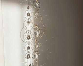 May include: A decorative hanging mobile with gold-colored rings, clear crystal spheres, and star accents. The mobile casts a shadow on a white wall, creating a play of light and shadow. The design includes various sizes of circles and dangling crystals.