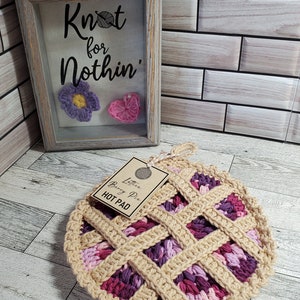 May include: Crocheted lattice berry pie hot pad in shades of pink and purple. The hot pad is shaped like a pie and has a brown paper tag that reads "Lattice Berry Pie Hot Pad".