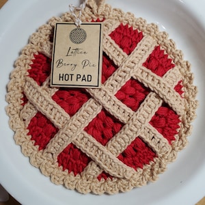 May include: A handmade, crochet hot pad in the shape of a lattice berry pie. The hot pad features a red center, tan lattice design, and a tan scalloped edge. A tag reads "Lattice Berry Pie HOT PAD".