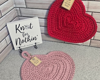 May include: Two crocheted heart-shaped hot pads, one red and one pink, with a brown tag that reads "Heart Hot Pad Trivet".