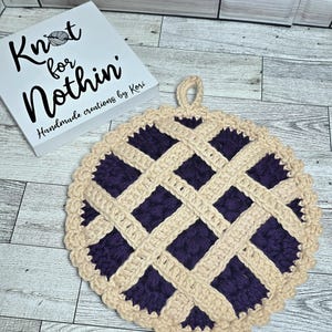 May include: A handmade crochet potholder in a pie design. The potholder features a dark purple filling with a beige lattice crust and edging. The potholder has a loop for hanging. A white sign with the text "Knot for Nothin" is in the background.
