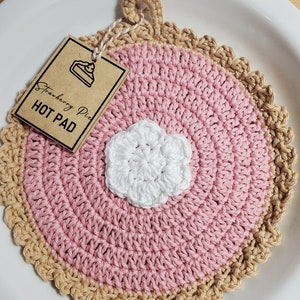May include: A pink and brown crocheted hot pad shaped like a pie with a white crocheted flower in the center. The hot pad has a brown tag that says "Strawberry Pie Hot Pad".