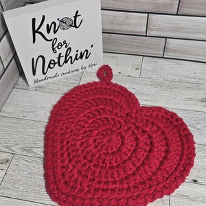 May include: A handmade, red crochet heart-shaped potholder. The potholder is made of thick yarn and has a loop for hanging. A white sign with the text "Knot for Nothin'" is in the background.